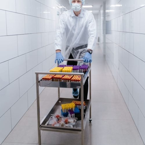 Lab technician in protective gear pushing a cart with test tubes in a sterile hallway.
